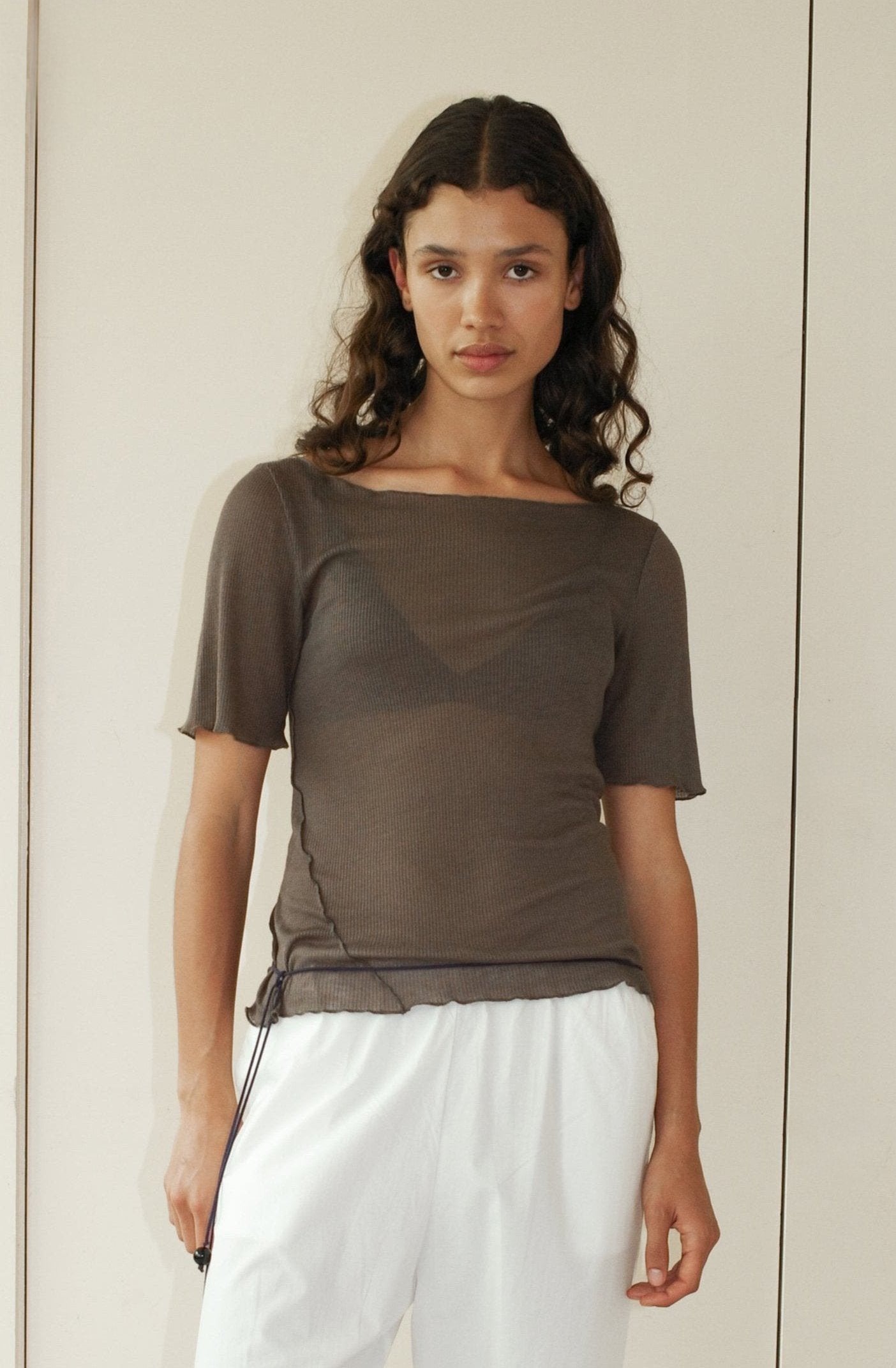 A young woman with wavy brown hair stands against a light wall, wearing the Square Knit Tee - Stone from Deiji Studios over a bralette and loose white pants, looking calmly at the camera.