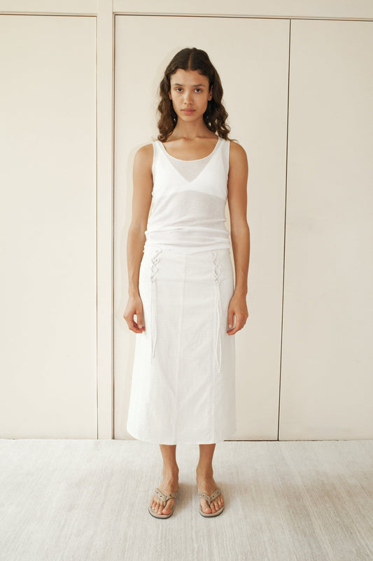 A woman stands indoors against light panels, wearing a Deiji Studios white sleeveless tank top, the Double Lace Up Skirt - Bone Check with side lace details, and beige sandals. She has long wavy hair and a neutral expression.