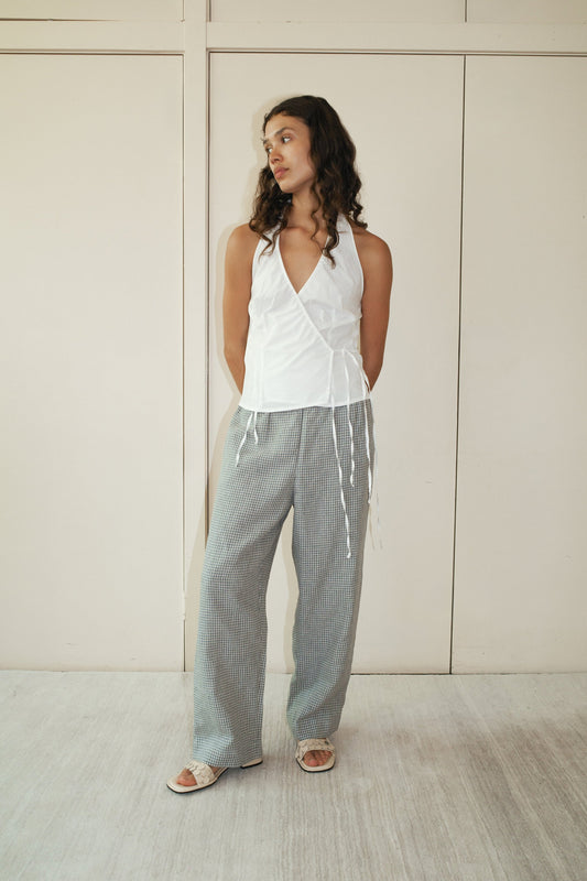 A woman with wavy hair stands against a beige wall, looking relaxed. She wears Deiji Studios’ Ease Trouser—loose gray checkered pants—paired with a sleeveless white wrap top and sandals.