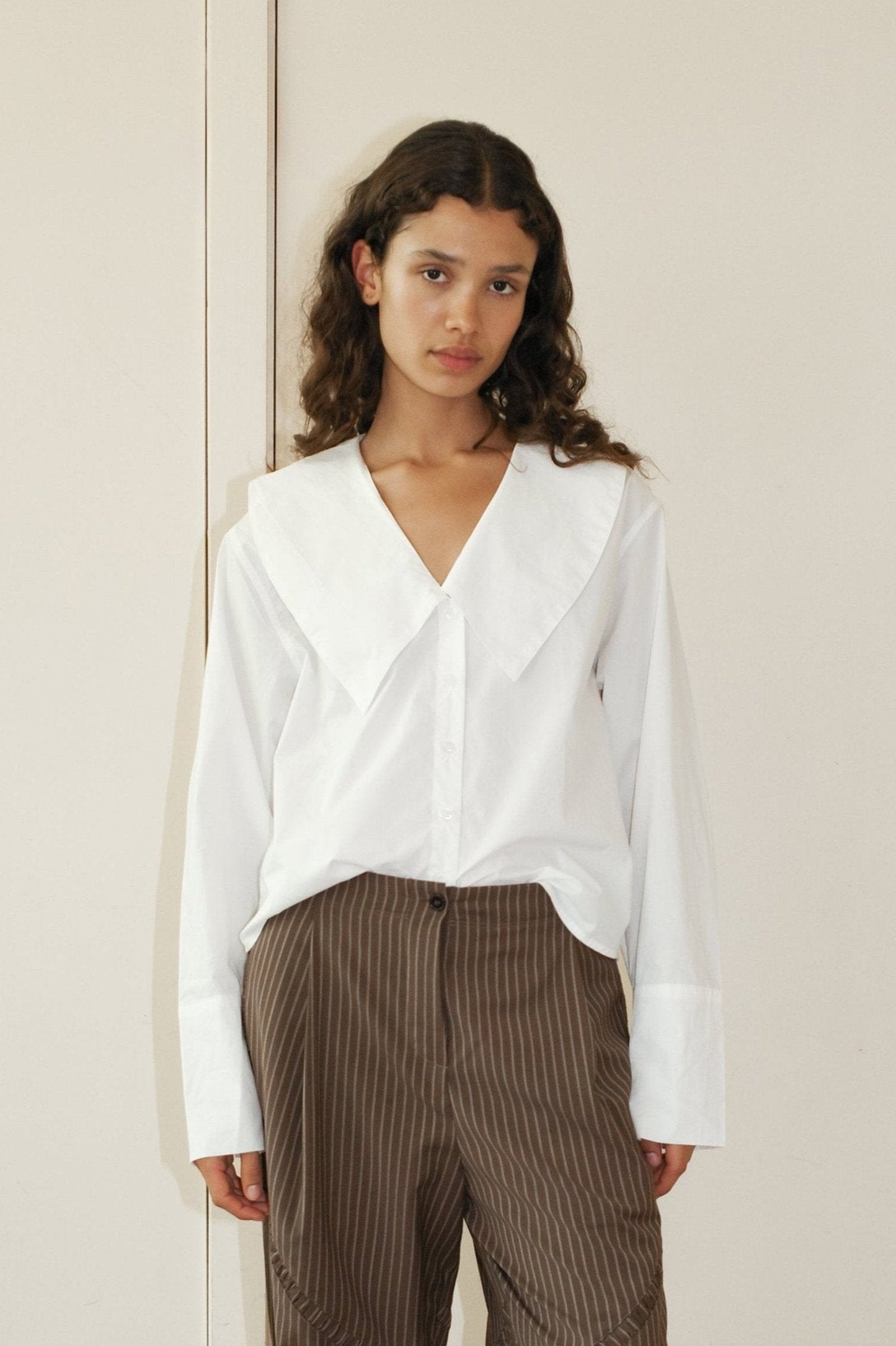 A young woman with long wavy hair and a neutral expression stands indoors against a light background, wearing the Oversized Collared Shirt in white with a large collar, paired with brown high-waisted pinstriped pants.
