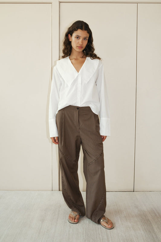 A woman with wavy dark hair stands indoors, wearing a white blouse with a large collar, 0 Channel Pant - Sable Stripe, and transparent sandals against a neutral wall background.