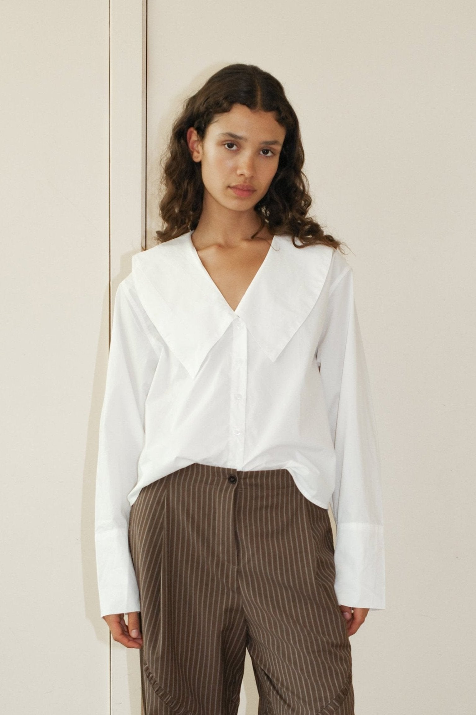 A young woman with long curly hair wears the Oversized Collared Shirt, a loose white blouse with a wide collar, and brown striped pants, standing against a cream background.