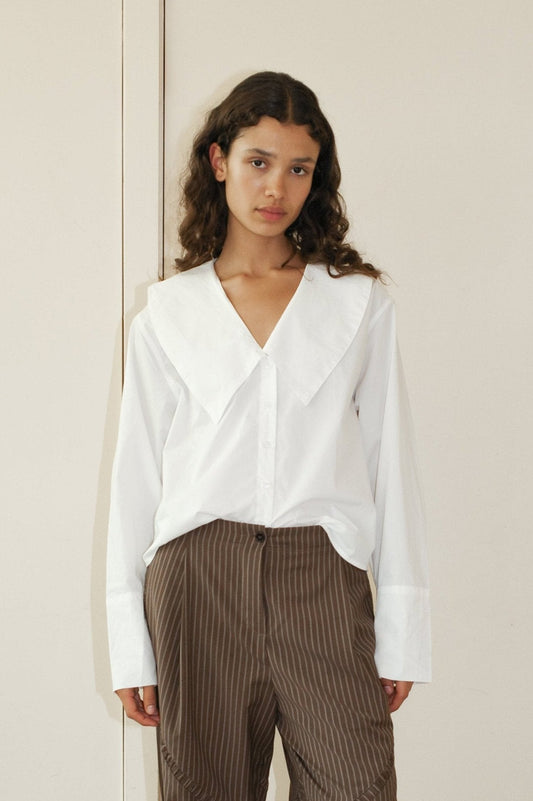 A young woman with long curly hair wears the Oversized Collared Shirt, a loose white blouse with a wide collar, and brown striped pants, standing against a cream background.