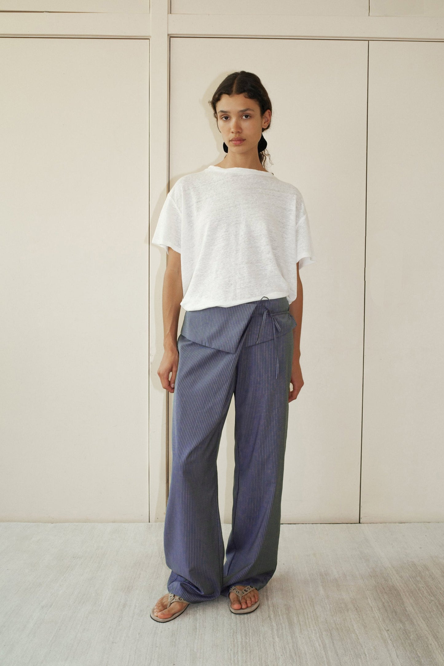 A person stands indoors against a light wall wearing 0s Folded Wool Pant in Navy Stripe, featuring relaxed straight legs and a wrap detail, paired with a loose white t-shirt and sandals.