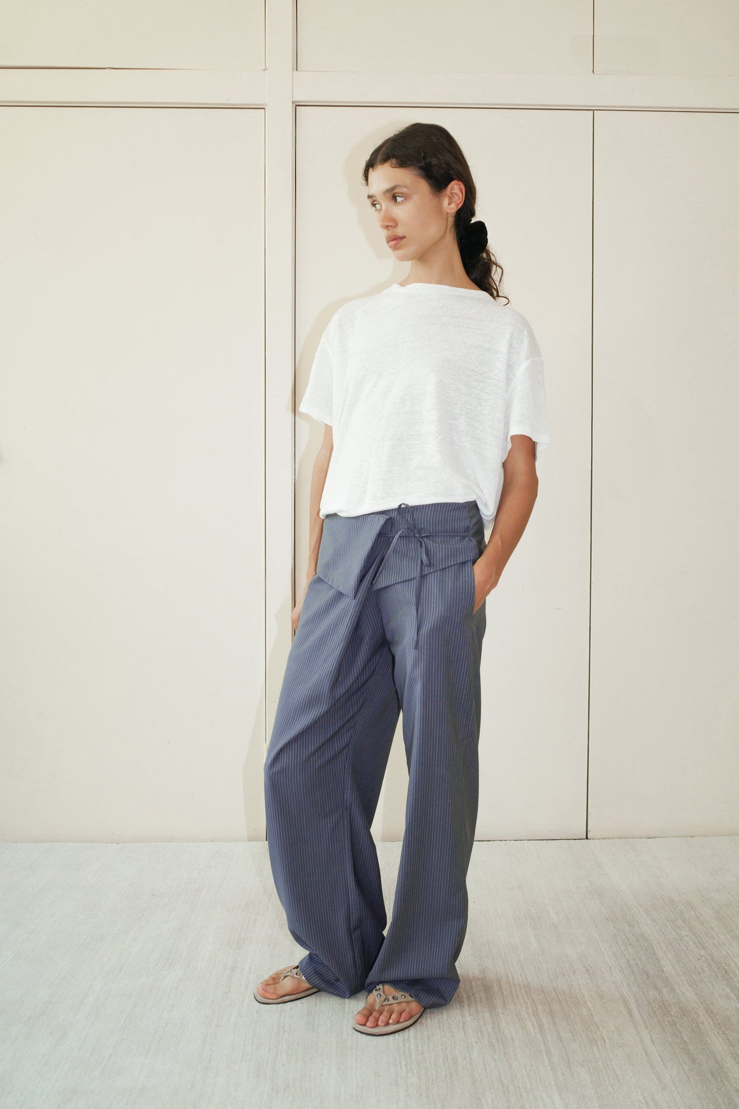 A woman stands indoors against a light wall, wearing a loose white t-shirt and 0 Folded Wool Pant - Navy Stripe with subtle stripes. She gazes to the side, hands in pockets, and finishes the look with sandals.