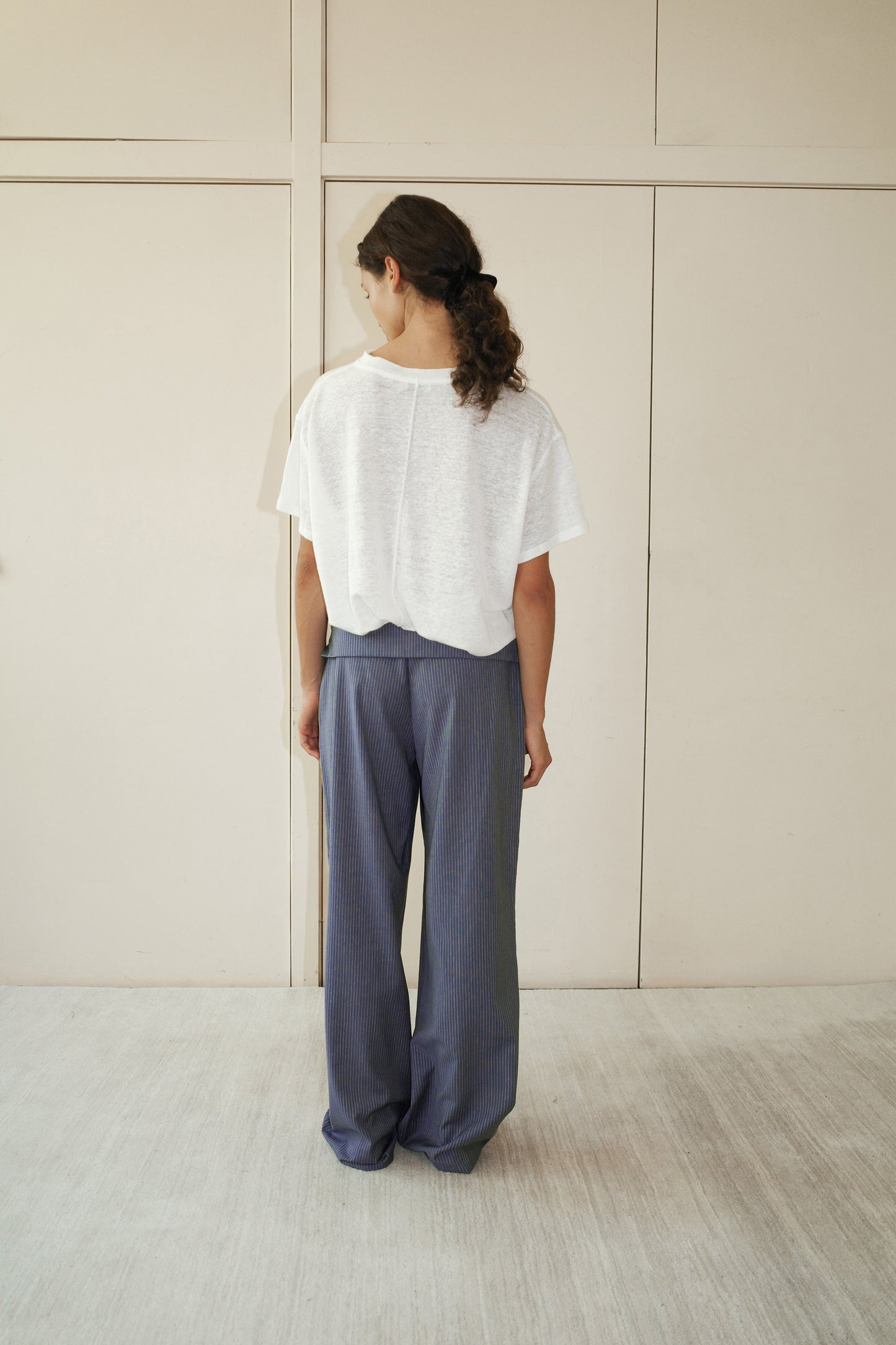 A person with long hair in a low ponytail stands facing away, wearing a loose white short-sleeve top and the 0 Folded Wool Pant - Navy Stripe, in a minimal indoor setting with light-colored walls and floor.