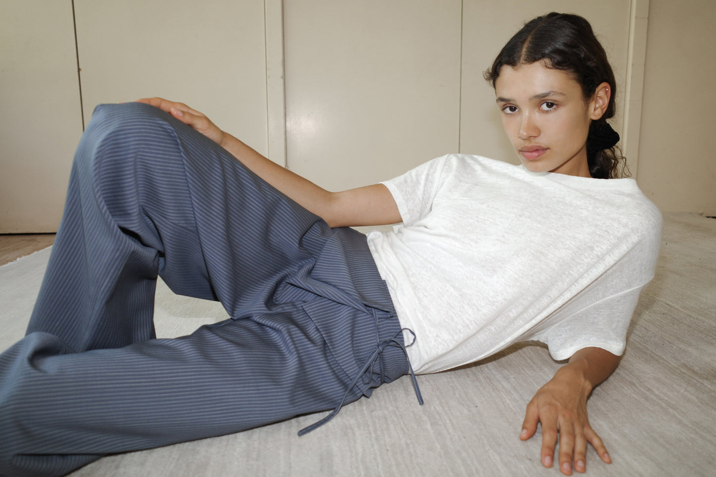 A person with long dark hair in a ponytail reclines on a light floor, wearing a loose white T-shirt and the Folded Wool Pant - Navy Stripe by 0, looking calmly at the camera.