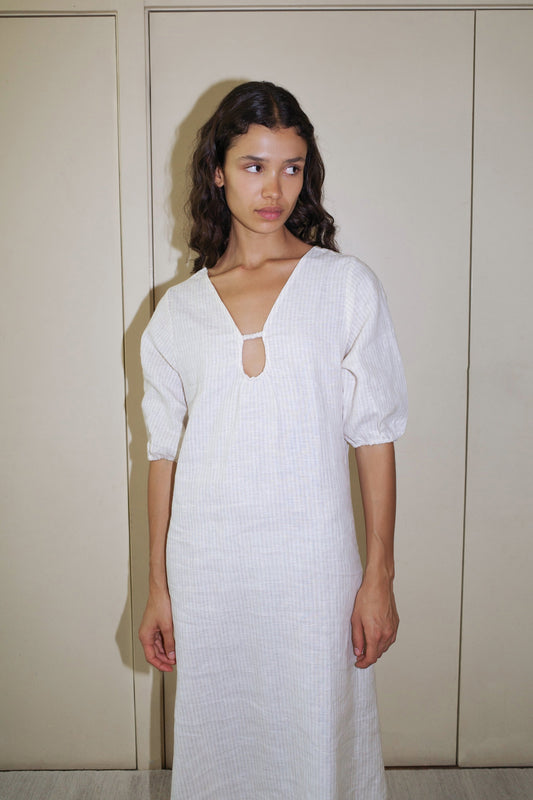 A woman with wavy dark hair wears the Squared Dress—a light-colored, long dress with half sleeves and a small front keyhole cutout—standing indoors against a beige wall, looking slightly to her left.