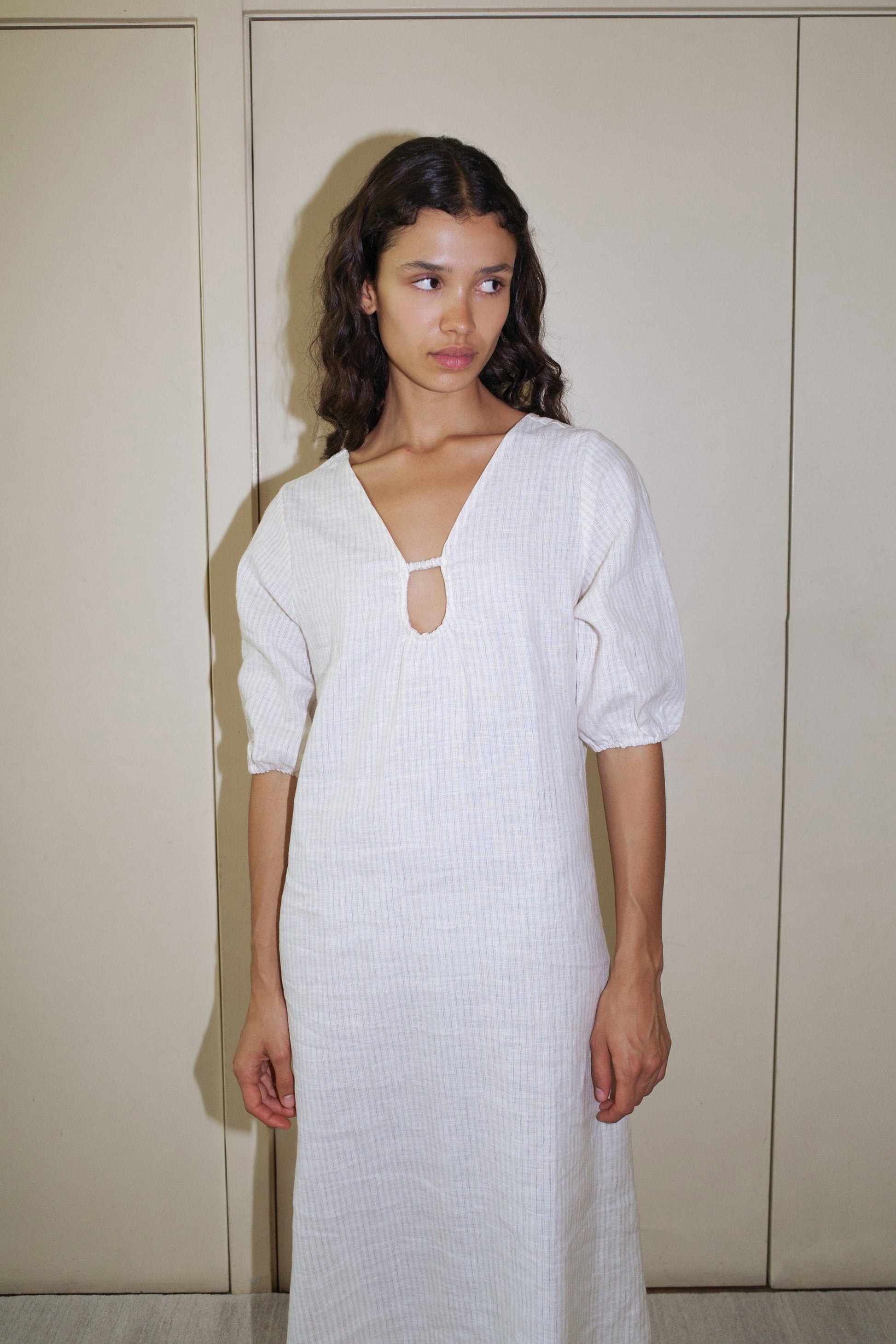 A woman with wavy dark hair wears the Squared Dress - Linen Story Stripe, a light-colored, knee-length dress in organic linen with elbow-length blouson sleeves and a keyhole neckline, standing against a beige wall looking to her left.