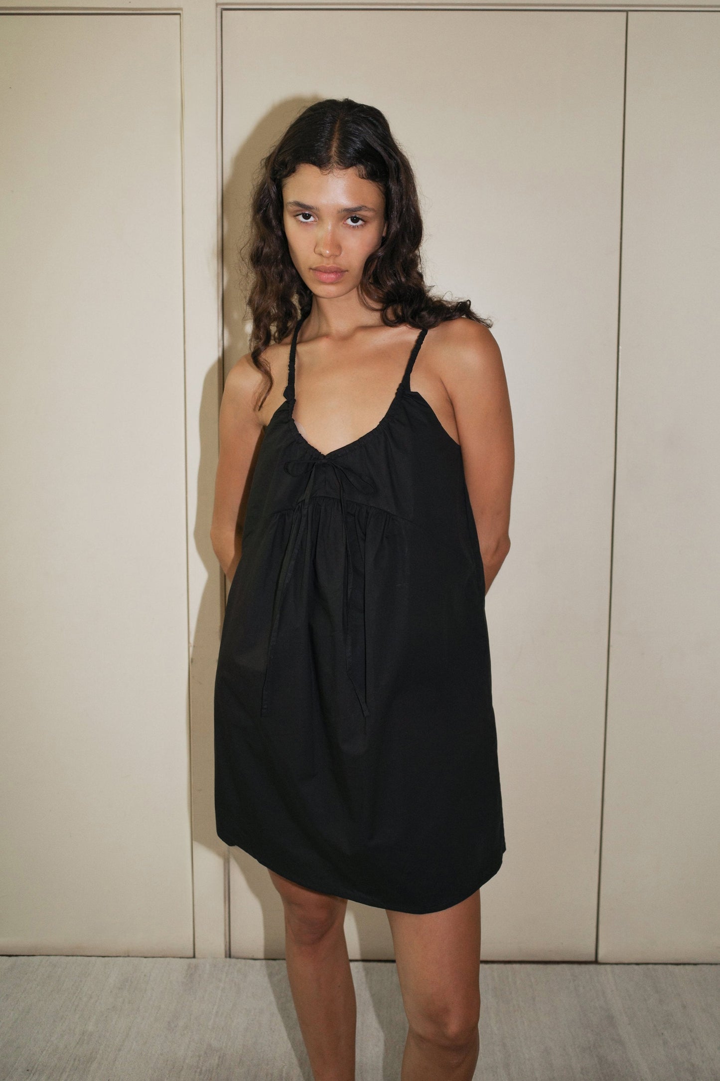 A woman with long, wavy hair stands indoors against a beige wall, wearing the Double V Dress—a loose black sleeveless dress featuring thin straps and a bow detail at the neckline.