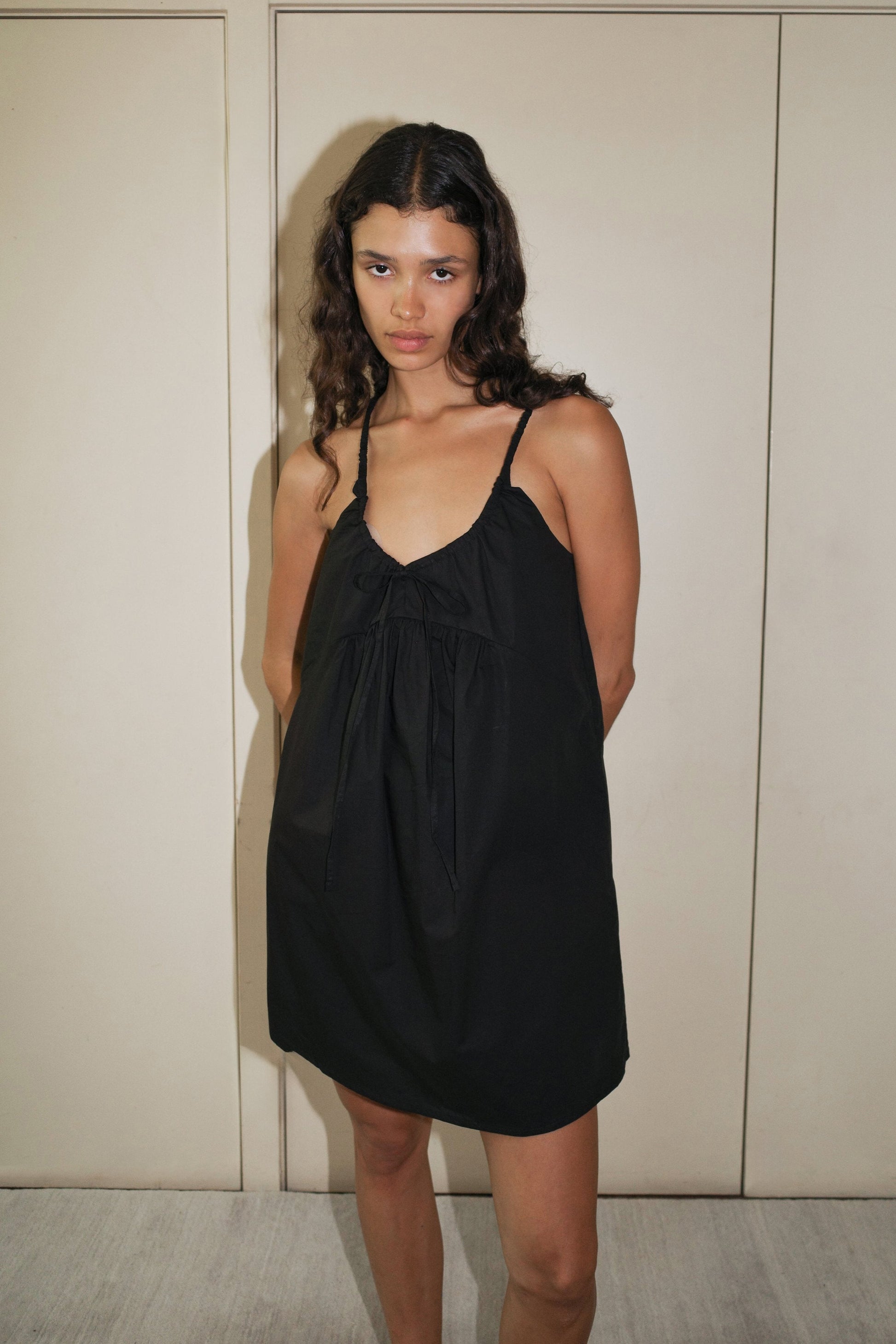 A woman with long, wavy hair stands indoors against a beige wall, wearing the Double V Dress—a loose black sleeveless dress featuring thin straps and a bow detail at the neckline.