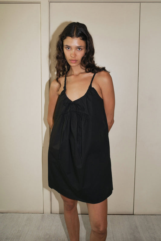 A woman with long, wavy hair stands indoors against a beige wall, wearing the Double V Dress—a loose black sleeveless dress featuring thin straps and a bow detail at the neckline.