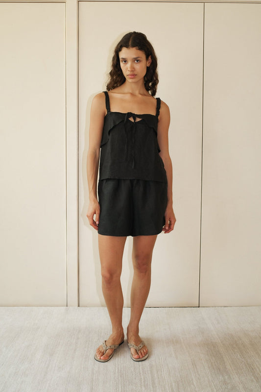 A woman stands indoors against a cream-colored wall, wearing the Tie Front Set—black sleeveless top with tie detail and matching shorts. She has wavy brown hair and beige flip-flops.