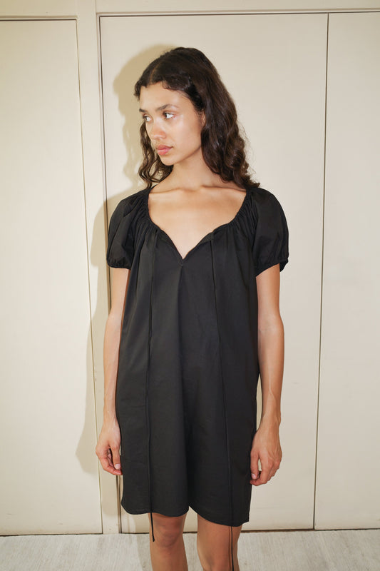 A woman with long, wavy dark hair wears the Capped Sleeve Dress, a short-sleeved black dress with a loose fit and deep V-neckline, standing indoors in front of closed beige doors and looking off to the side.
