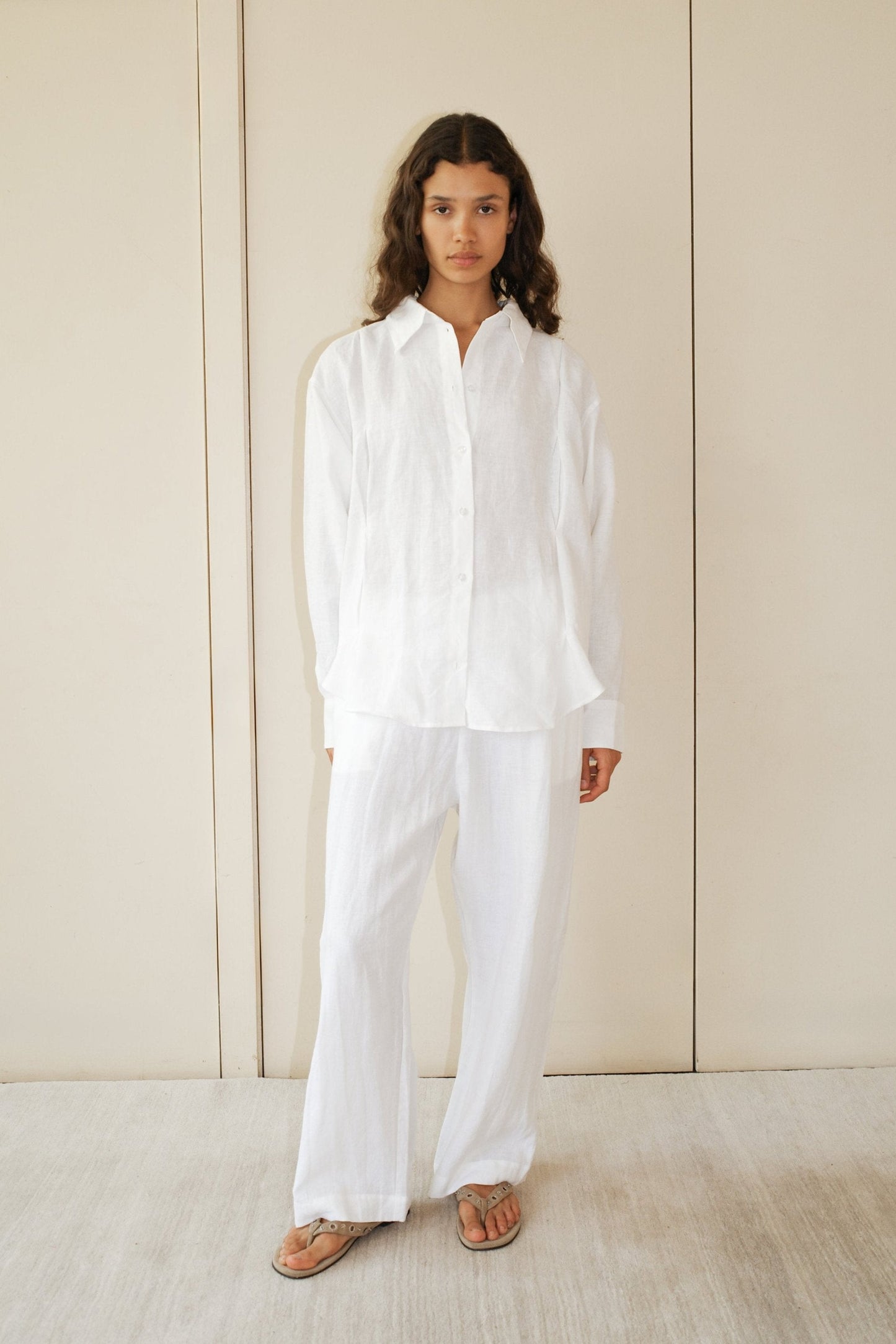 A person with long, wavy hair stands indoors against a neutral background, wearing the Tack Set—an airy white button-up shirt and matching pants—paired with tan sandals.