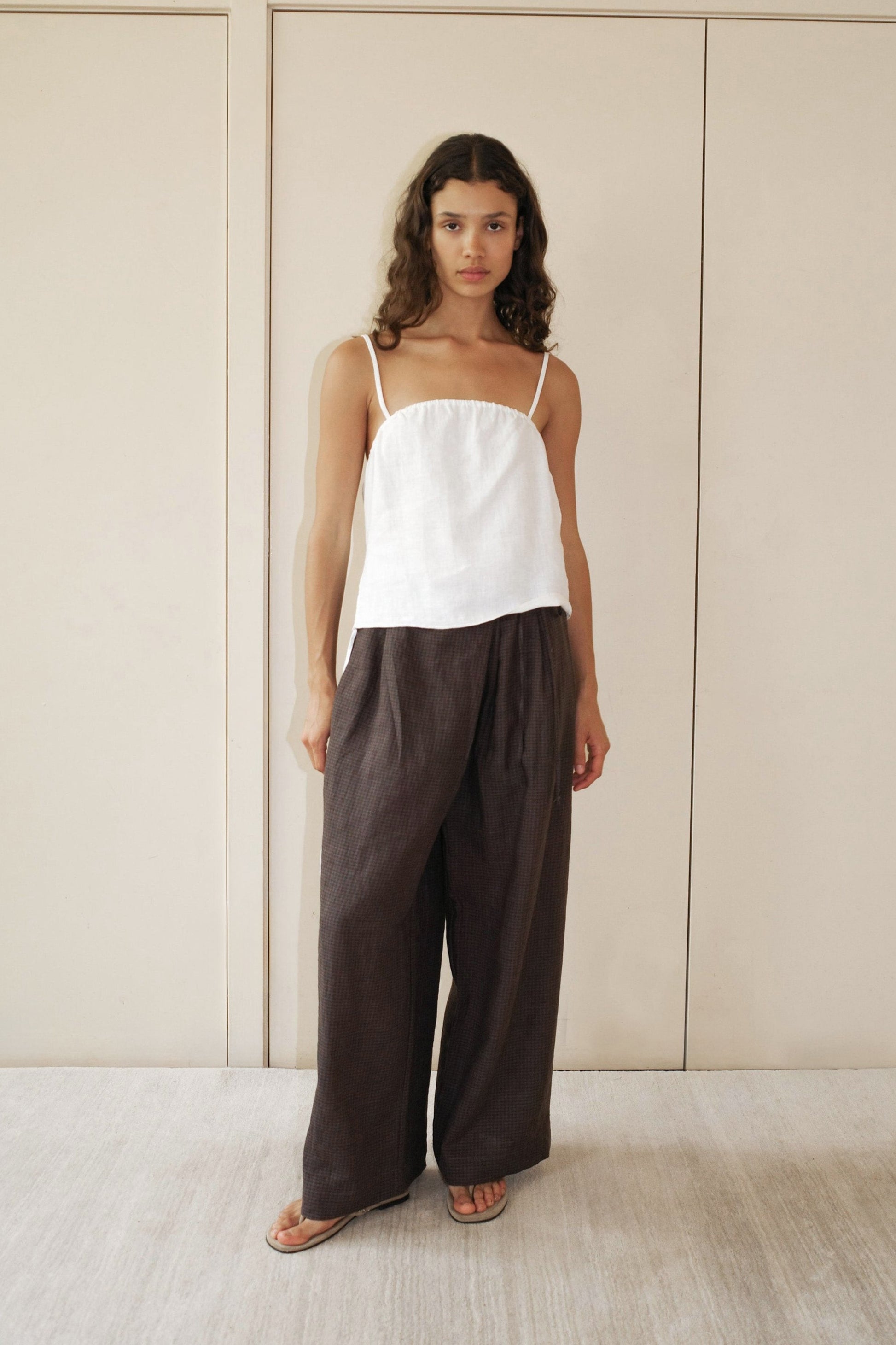 A woman with long curly hair wears the Babylock Linen Top - White with thin straps and loose, wide-legged dark brown pants, standing barefoot on a light floor before beige paneled doors.