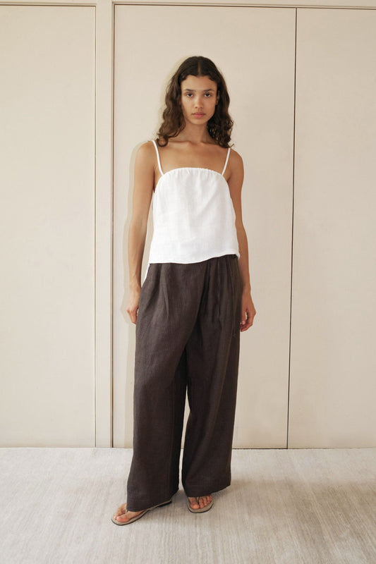A woman with long curly hair wears the Babylock Linen Top - White with thin straps and loose, wide-legged dark brown pants, standing barefoot on a light floor before beige paneled doors.