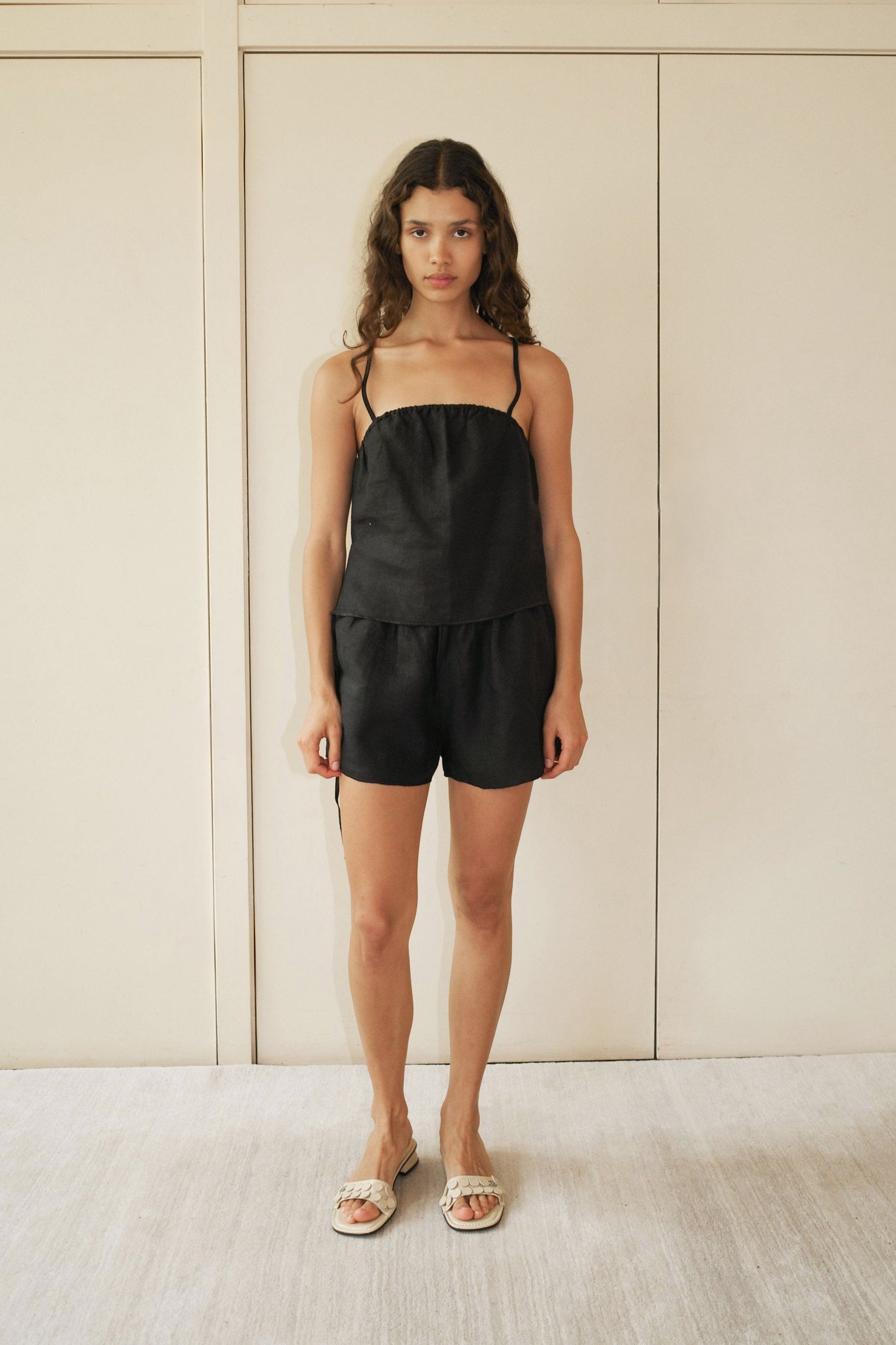 A woman with medium-length wavy hair stands indoors against a beige background, wearing the Babylock Linen Set - Black, which features a black linen top with thin straps and matching black shorts. She completes the look with white sandals.