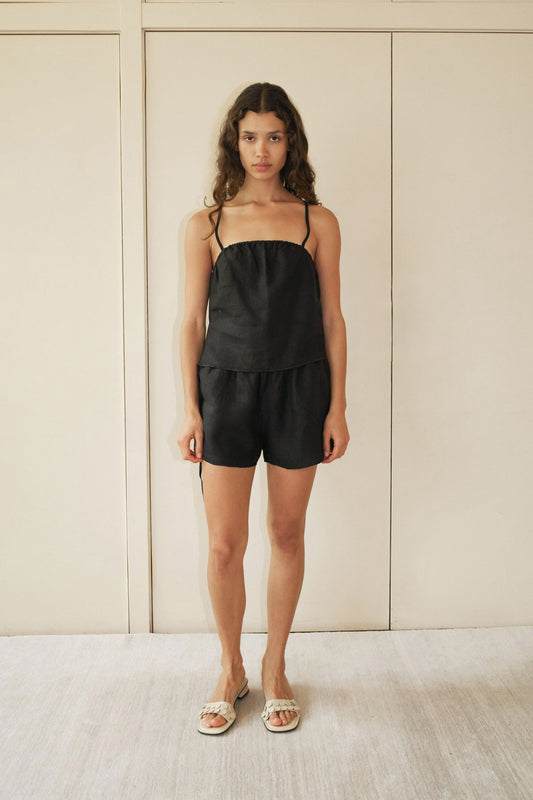 A woman with medium-length wavy hair stands indoors against a beige background, wearing the Babylock Linen Set - Black, which features a black linen top with thin straps and matching black shorts. She completes the look with white sandals.
