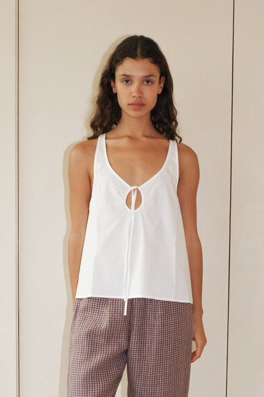 A person with long curly hair wears the U Shape Top, a sleeveless white top with a keyhole cutout and neckline tie, paired with loose brown plaid pants, standing against a plain beige background.