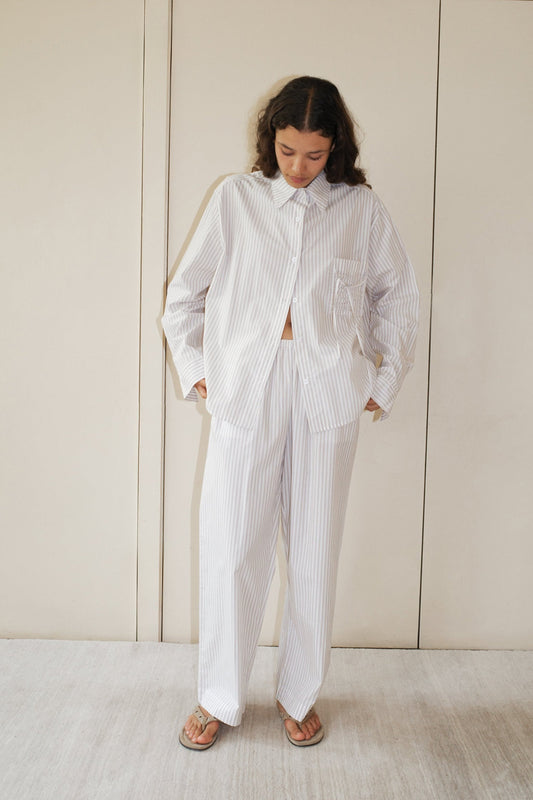 A person with long curly hair stands indoors looking down, wearing the Deiji Studios Pocket Set - Lake Stripe, a relaxed fit, white organic cotton poplin button-up shirt and matching wide-leg pants with light vertical stripes, paired with flip-flops.