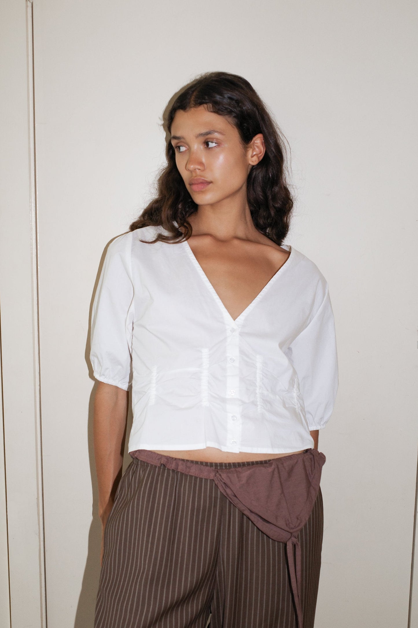 A woman with long, wavy dark hair wears the Deiji Studios Draw Top - White in organic cotton poplin and brown pinstriped pants. She stands against a plain light wall, looking to her left with a neutral expression.