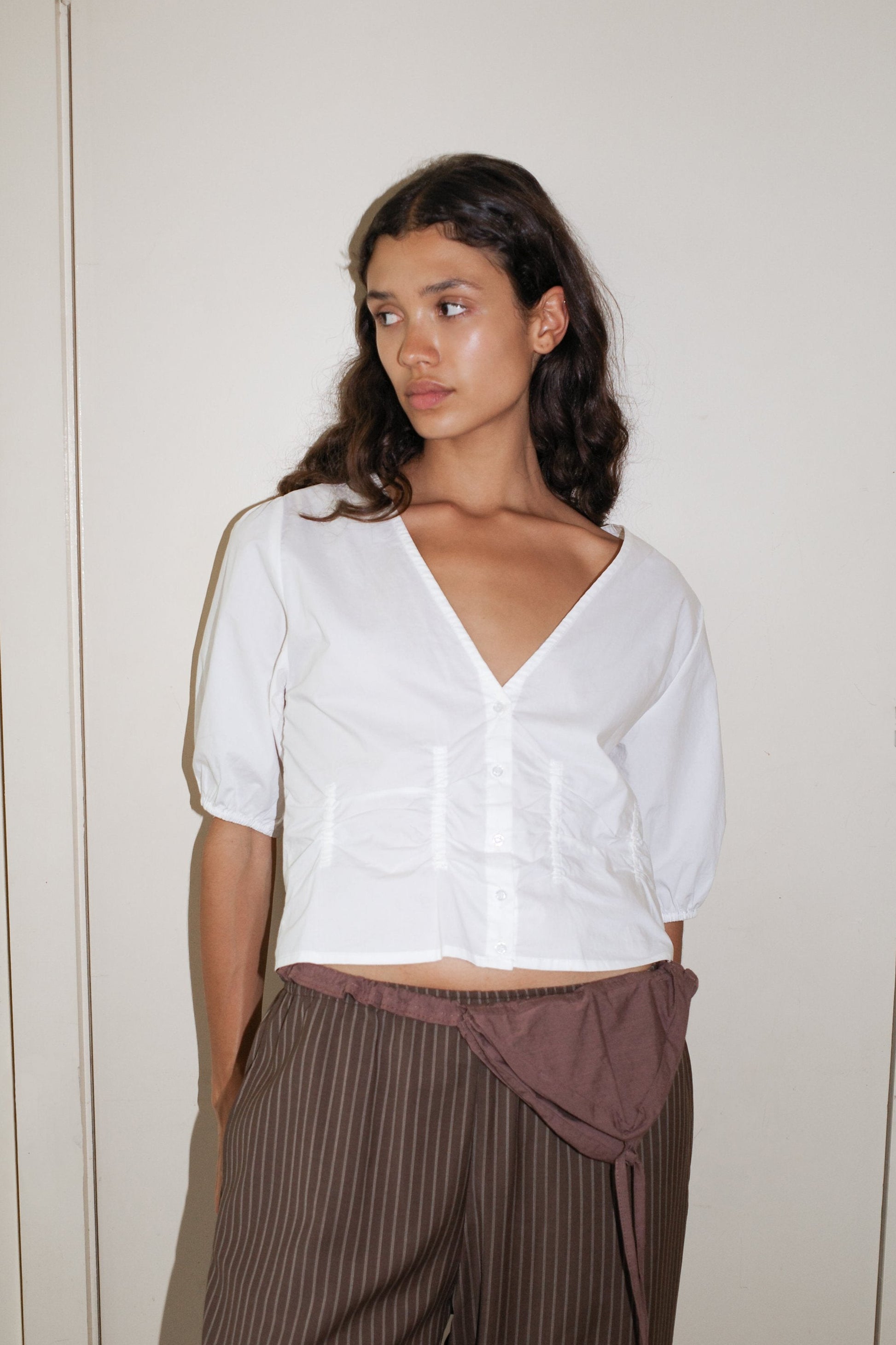 A woman with long, wavy dark hair wears the Deiji Studios Draw Top - White in organic cotton poplin and brown pinstriped pants. She stands against a plain light wall, looking to her left with a neutral expression.