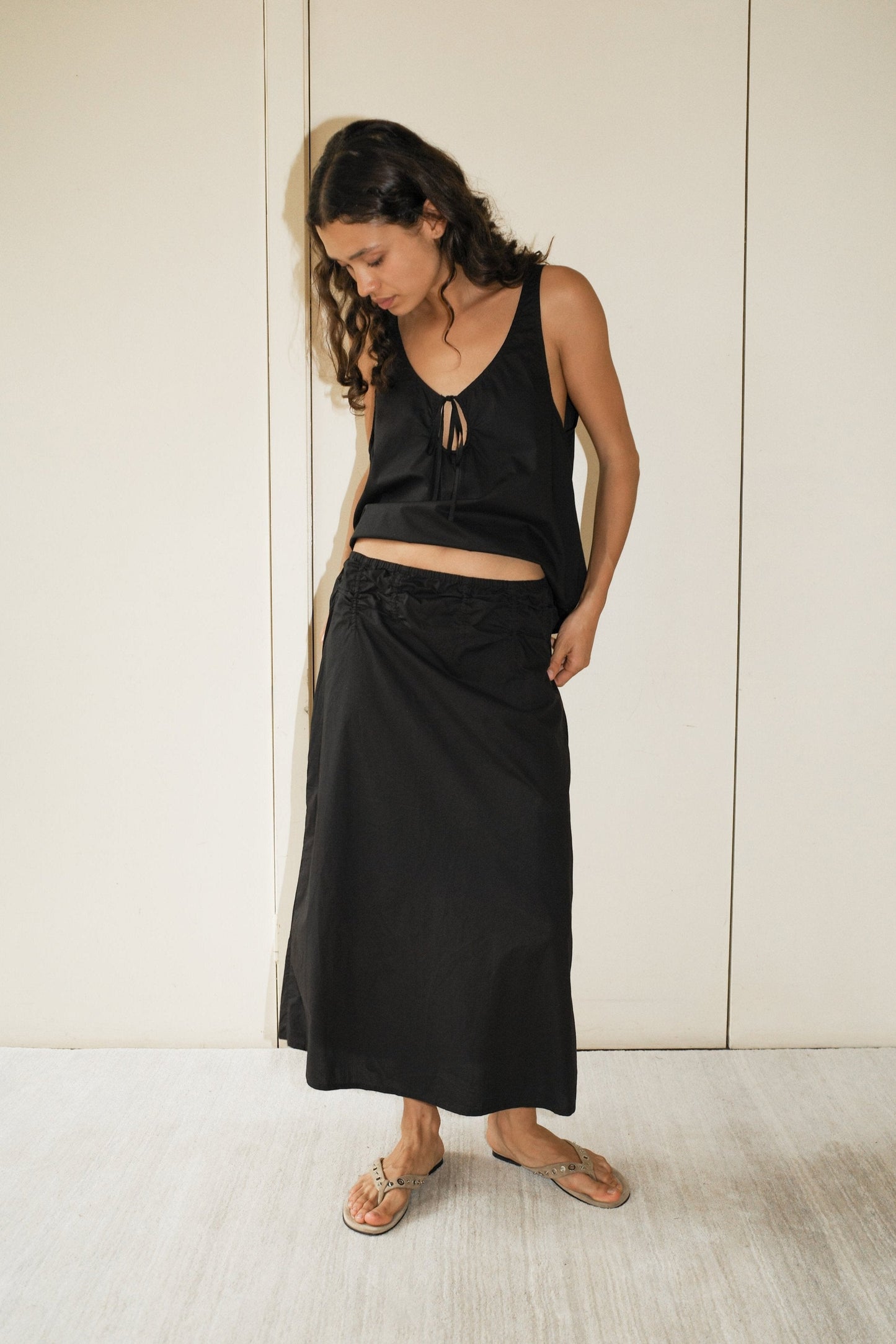 A woman with long curly hair stands indoors, barefoot on a light floor, wearing a sleeveless black top and the Channel Skirt - Black, an organic cotton midi skirt with an elastic waist. She looks down while holding her skirt.