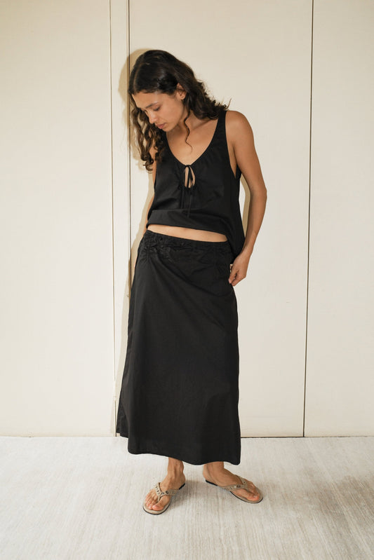 A woman with long curly hair stands indoors, barefoot on a light floor, wearing a sleeveless black top and the Channel Skirt - Black, an organic cotton midi skirt with an elastic waist. She looks down while holding her skirt.