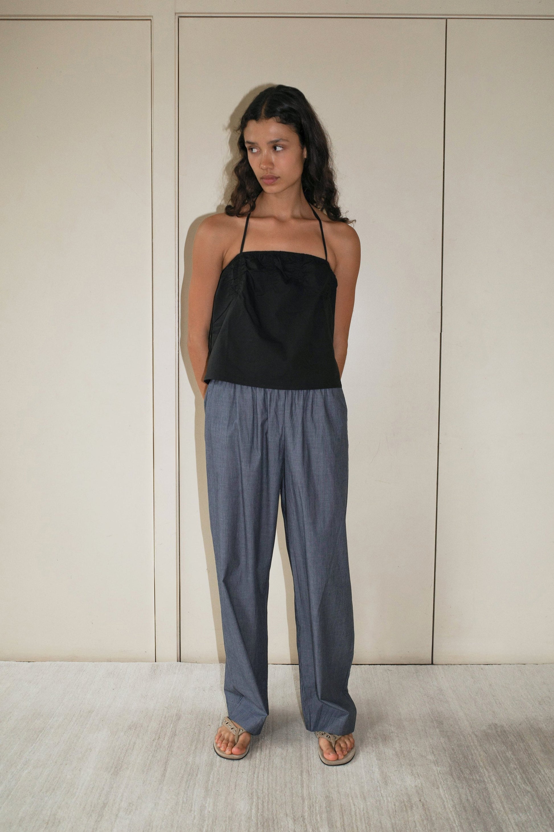A woman stands indoors against beige panel doors, wearing the Ease Trouser by Deiji Studios with a black halter-neck top and tan sandals. Her arms are behind her back as she gazes to the side with a neutral expression.