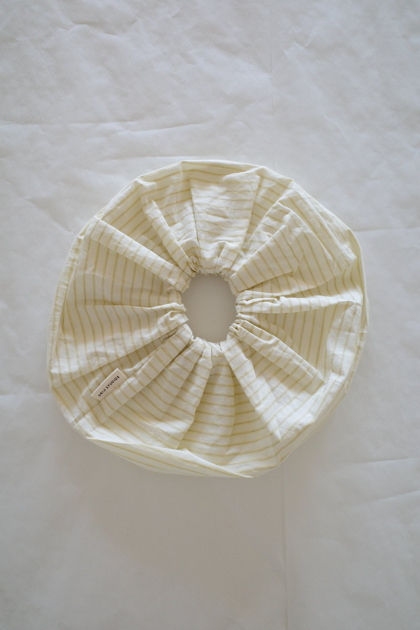 A Scrunchie with yellow stripes, made of fabric and featuring an elastic edge, lies flat on a white surface. A small fabric tag is attached to it.