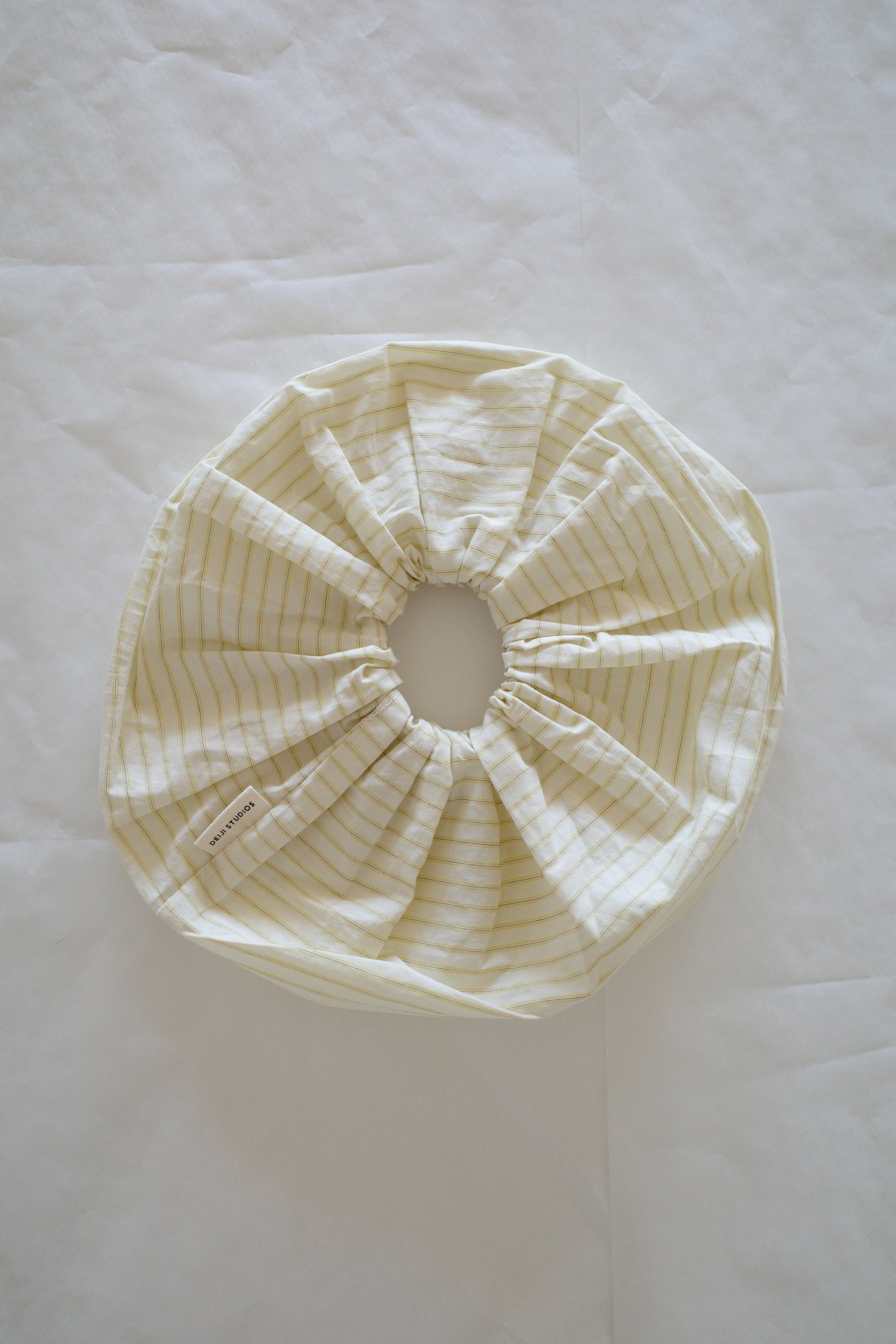 A Scrunchie with yellow stripes, made of fabric and featuring an elastic edge, lies flat on a white surface. A small fabric tag is attached to it.