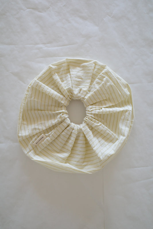 A round, off-white Scrunchie with yellow stripes and a gathered elastic center, resembling a shower cap, is laid flat on a white surface, featuring a small fabric tag on one side.