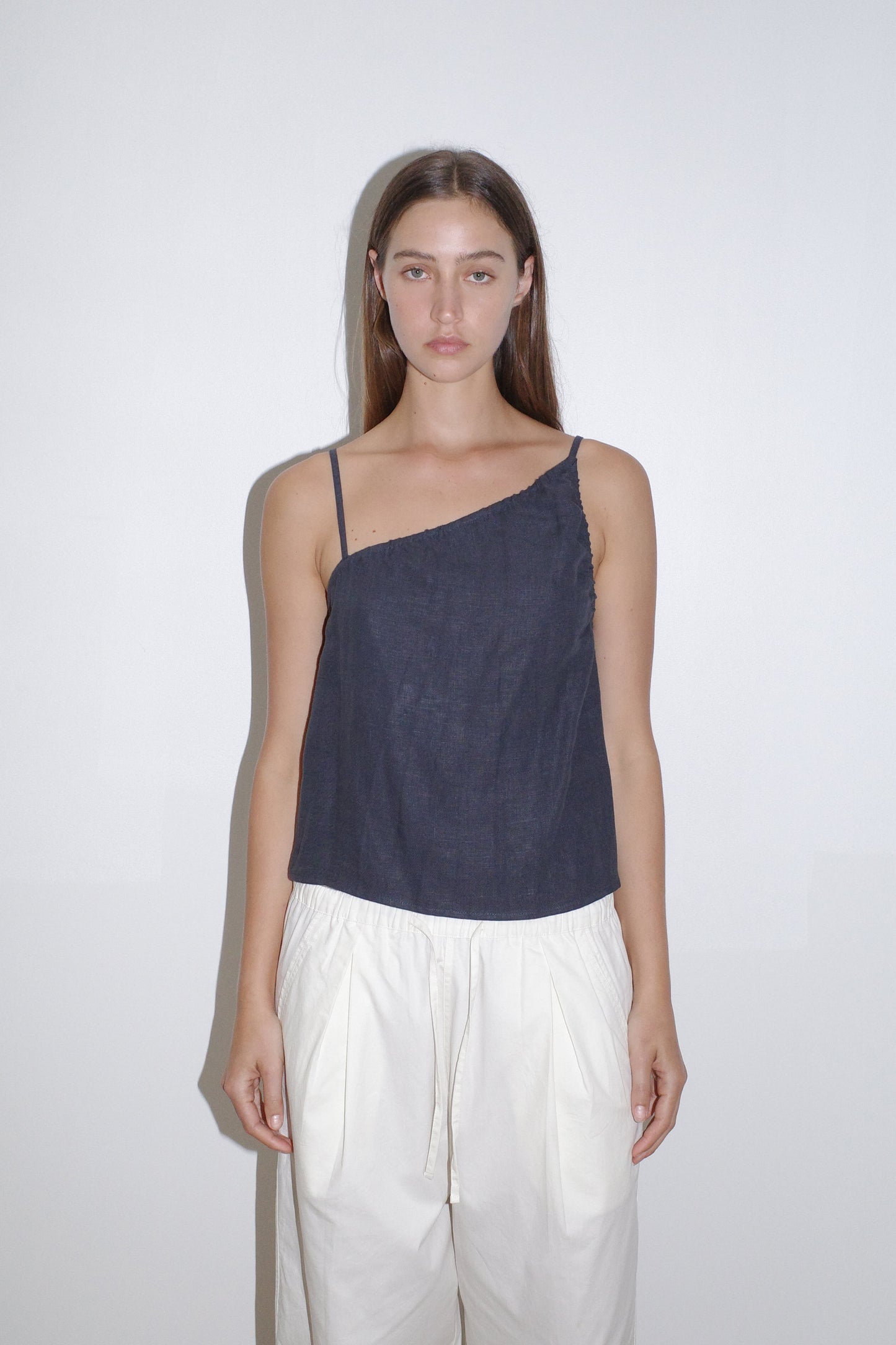A woman with long brown hair stands against a white wall, wearing the navy Asymmetric Tie Top and loose white pants. She has a neutral expression and her arms rest at her sides.