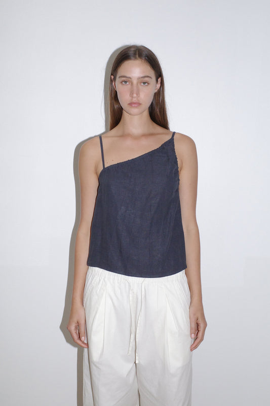 A woman with straight brown hair stands against a white wall, wearing the Asymmetric Tie Top in navy blue paired with loose white pants. She has a neutral expression and her arms rest by her sides.