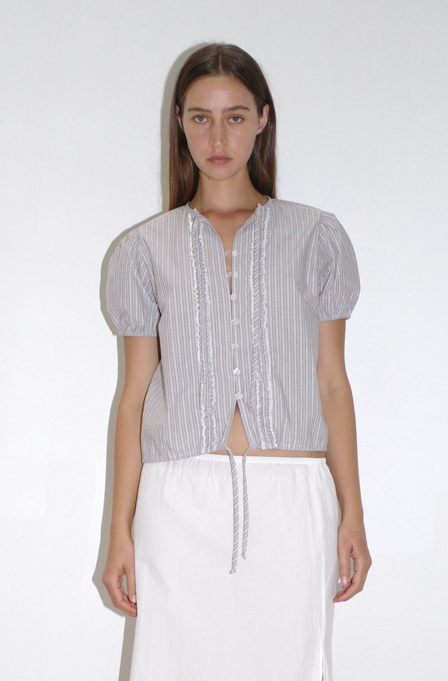 A woman with straight brown hair and a neutral expression stands against a plain light background, wearing the Deiji Studios Double Frill Top - Seas Stripe with ruffles and a tie at the waist, paired with a long white skirt.