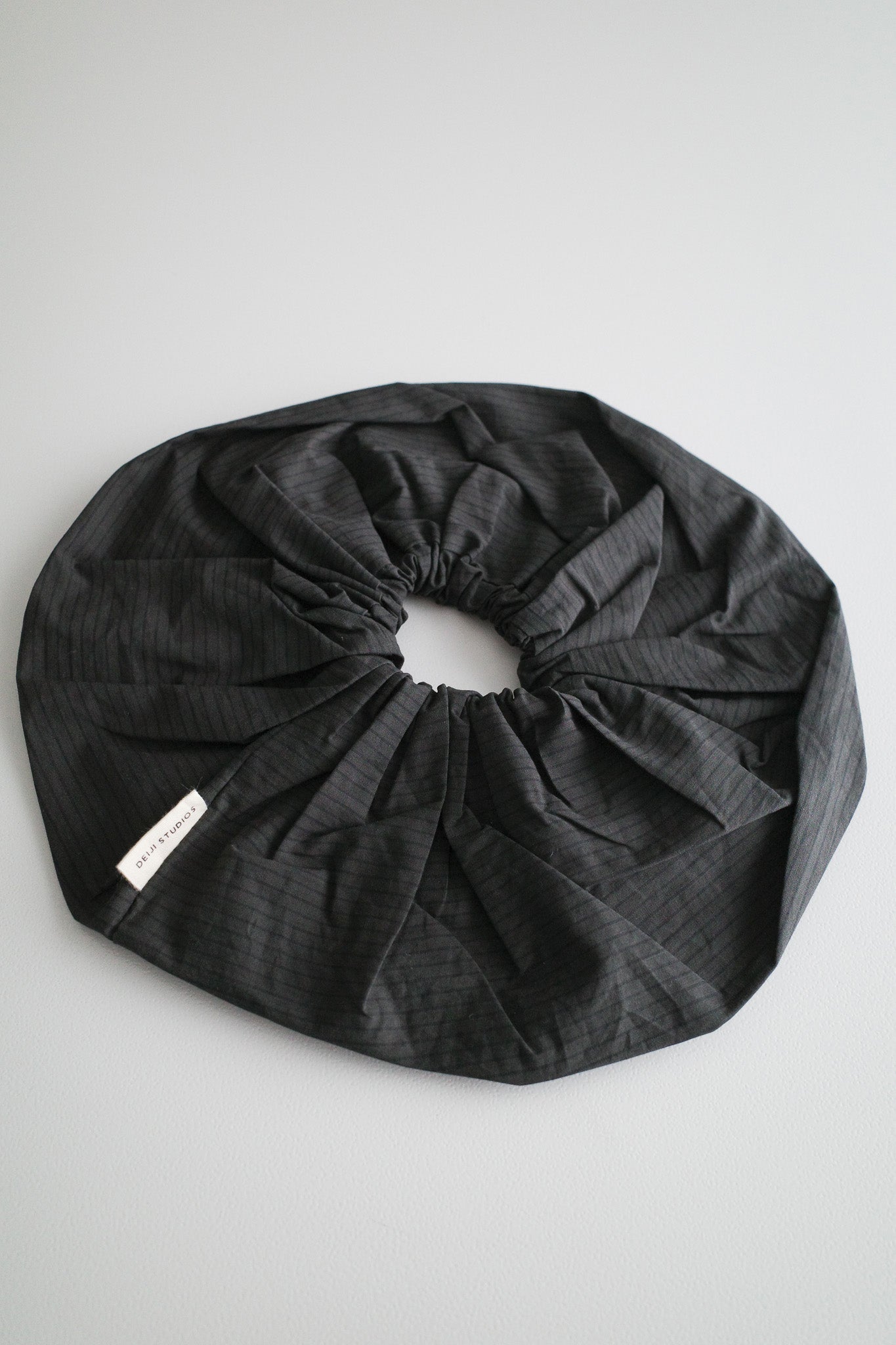 A large, black, pinstriped oversized scrunchie with a fabric tag attached—Scrunchie - Black Stripe by Deiji Studios—displayed on a light gray surface.