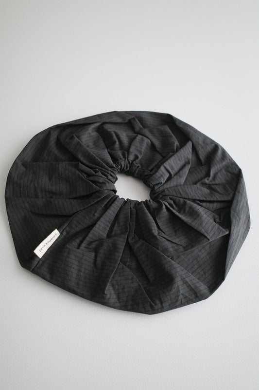 The Scrunchie - Black Stripe is a large, black oversized accessory with subtle pinstripes, made from soft organic cotton. Displayed flat on a light gray surface, it features a small white text tag on the edge.