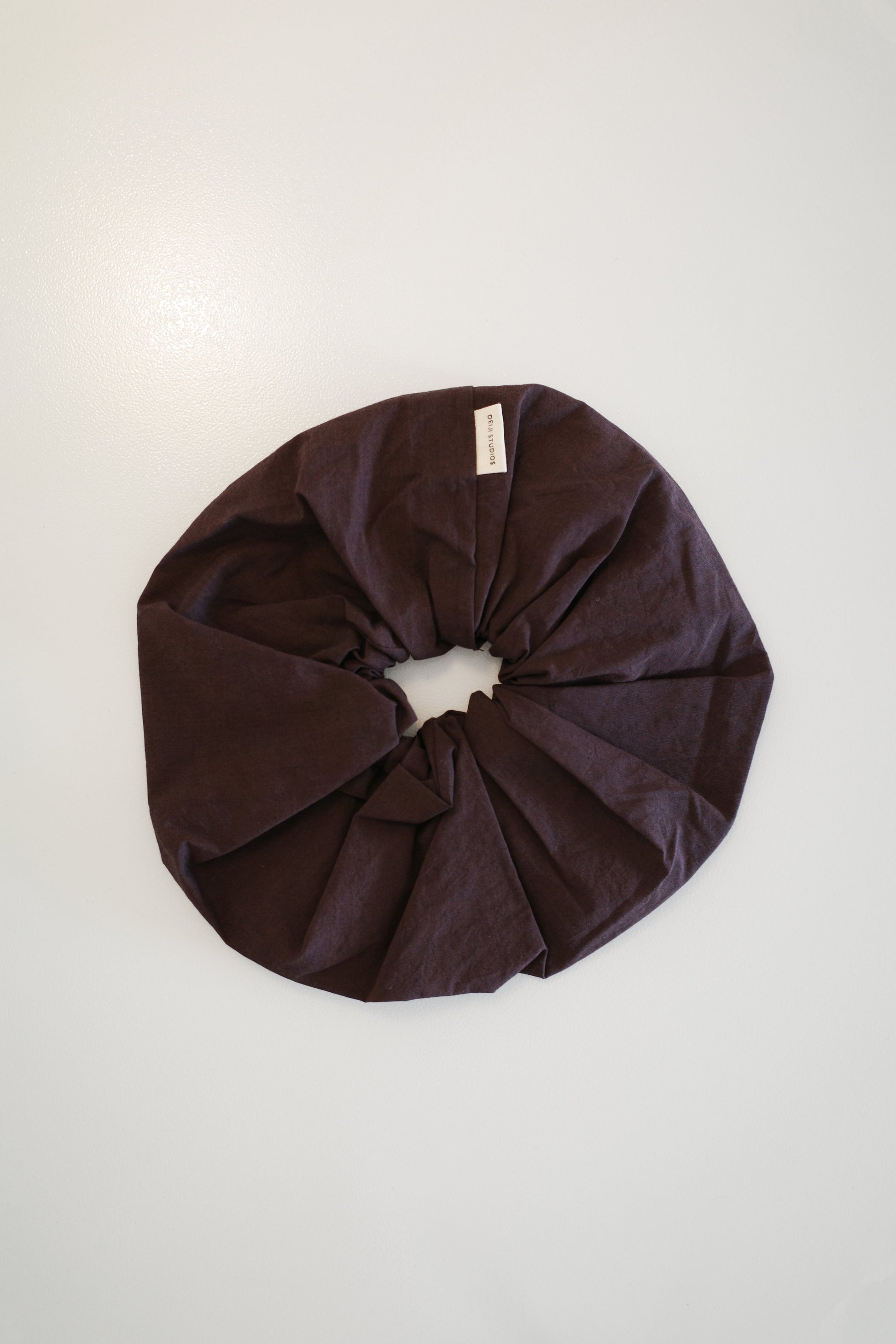 The Scrunchie - Umber from Deiji Studios, a large dark brown oversized scrunchie with a small white tag, is displayed in a circle on a plain white background.