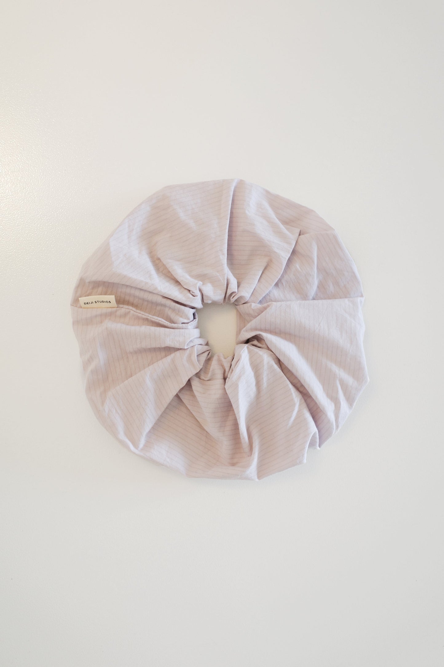 A large, light lavender Scrunchie with subtle stripes is laid flat on a white surface, featuring a small beige tag sewn into its side.
