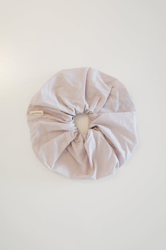 A large, light lavender Scrunchie with subtle stripes is laid flat on a white surface, featuring a small beige tag sewn into its side.