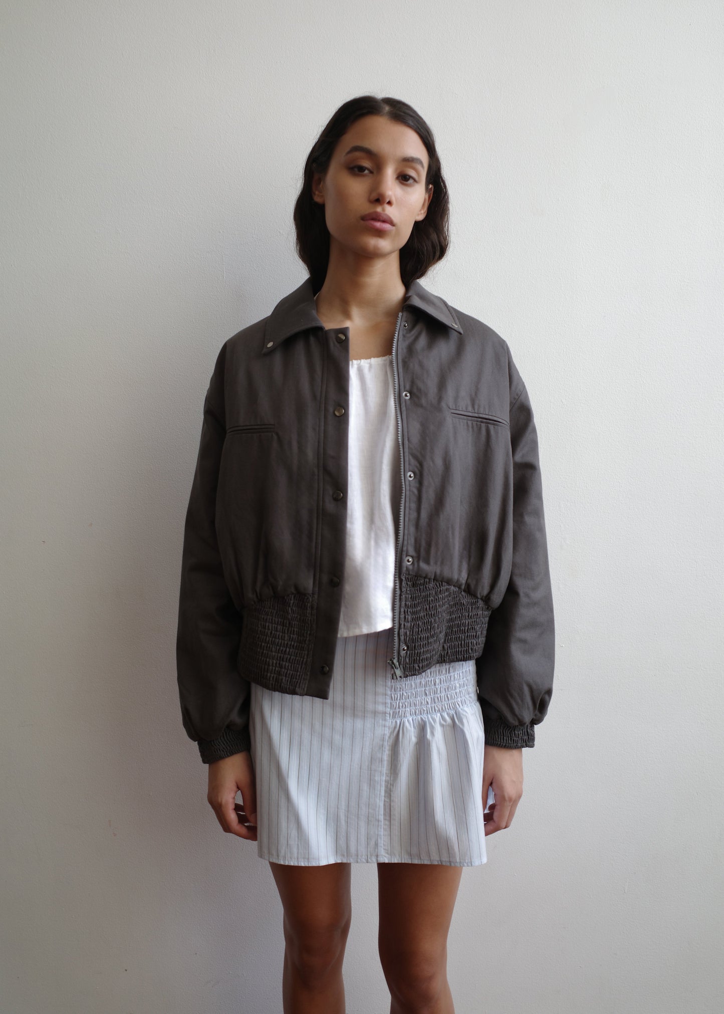 A woman with long dark hair stands against a white wall, wearing a loose gray jacket over a white top and the Shirring Skirt - Mineral Stripe from Deiji Studios. She has a neutral expression and her arms rest at her sides.