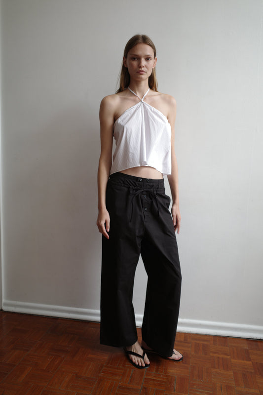 A woman stands indoors against a white wall, wearing a sleeveless white halter top, Deiji Studios Two Band Pant - Black tied at the waist, and black sandals. She has straight hair and a neutral expression.
