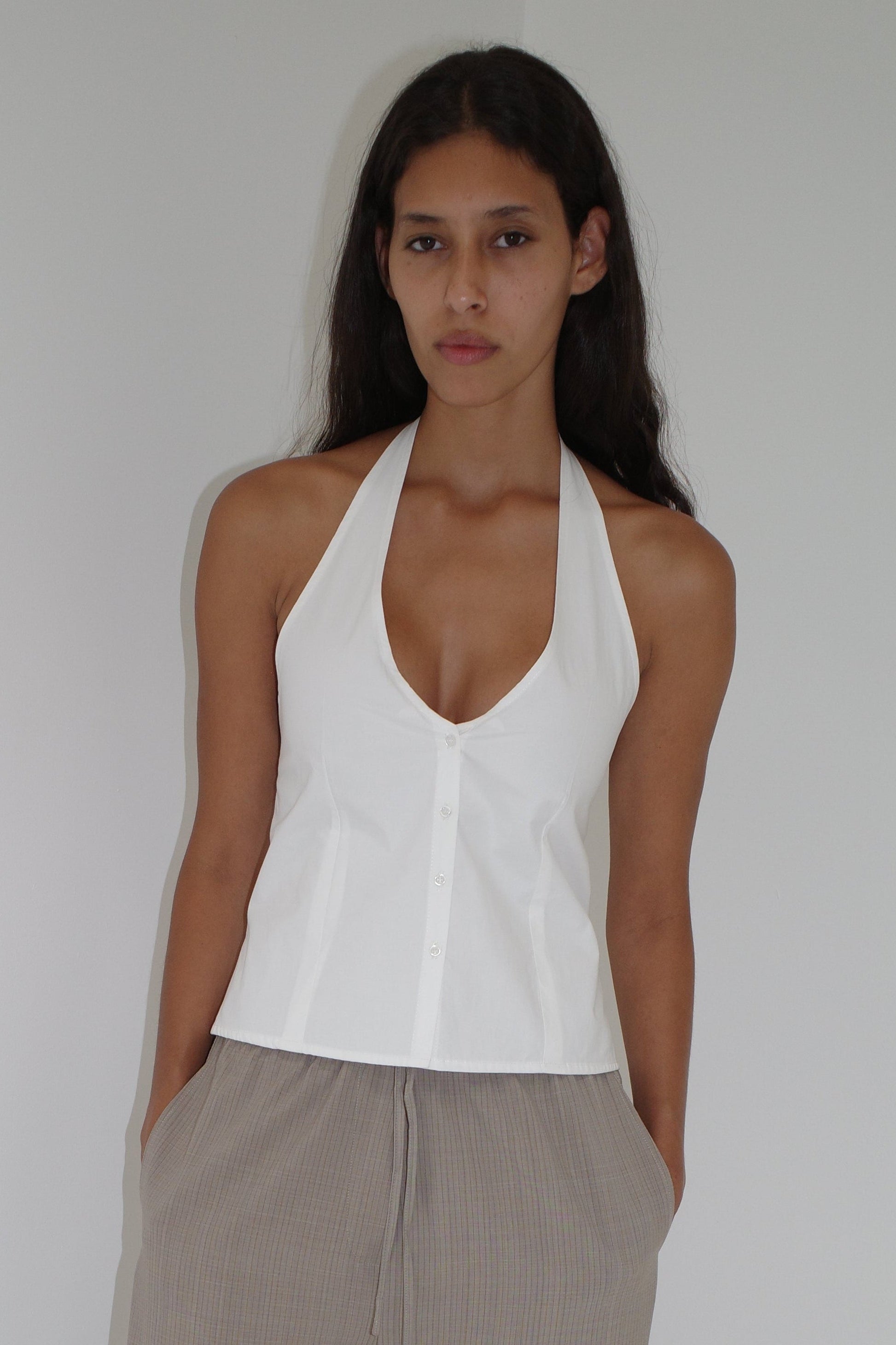 A woman with long dark hair stands against a plain wall, wearing a sleeveless white Waist Halter top and light brown pleated pants, her hands in her pockets and a neutral expression.