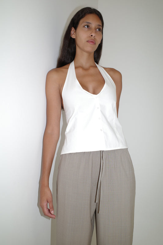 A woman with long dark hair and a neutral expression stands against a white wall, wearing the Low Pocket Pure Wool Pant in Dove Stripe and a white halter-neck top with buttons. The high-waisted pants have a relaxed fit and drawstring detail.