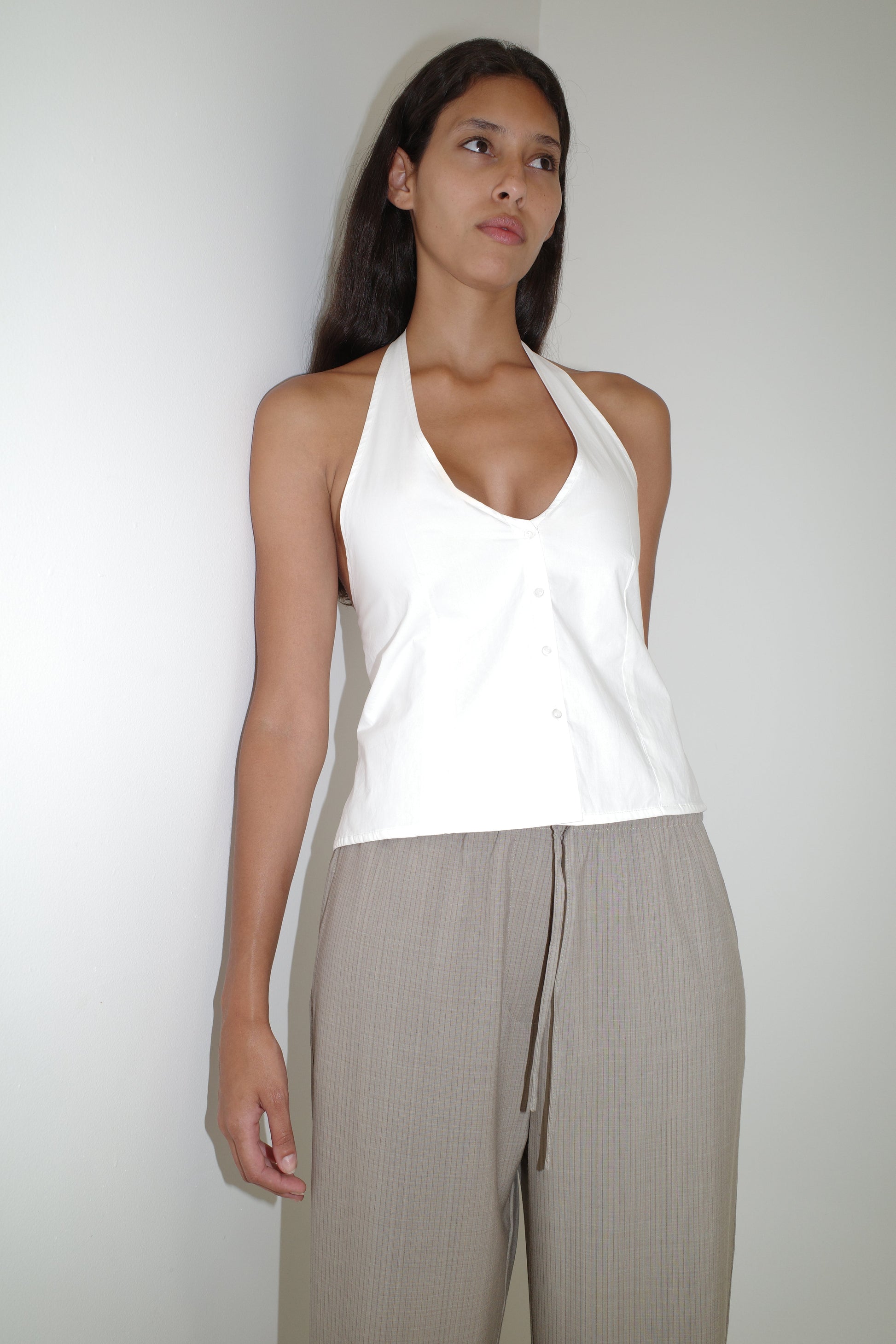 A woman with long dark hair and a neutral expression stands against a white wall, wearing the Low Pocket Pure Wool Pant in Dove Stripe and a white halter-neck top with buttons. The high-waisted pants have a relaxed fit and drawstring detail.