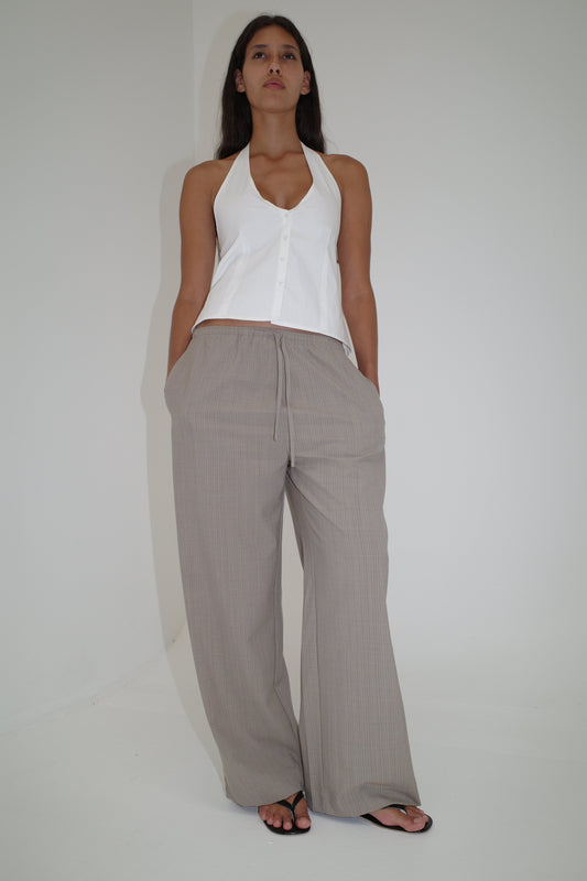 A woman stands against a plain white wall, wearing a sleeveless halter top, black sandals, and the Low Pocket Pure Wool Pant in Dove Stripe. She looks confidently at the camera with her hands in her pockets.