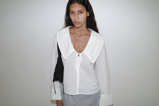 A woman with long dark hair stands against a white wall, wearing the Collared Tie Shirt - White, a light gray skirt, and carrying a black shoulder bag. She looks directly at the camera with a neutral expression.