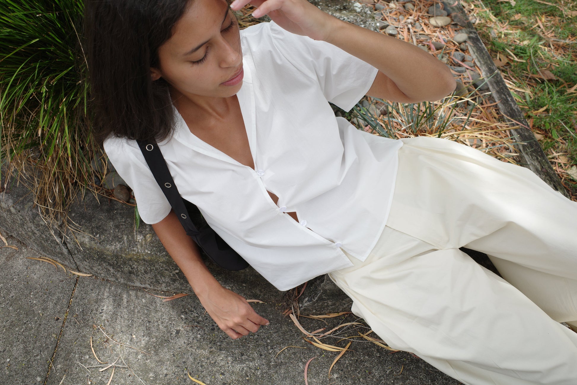 A person with long dark hair, dressed in the Sails Top - White from Deiji Studios and light pants, sits on a curb outdoors amid dry leaves and grass, looking down with one hand near their face.