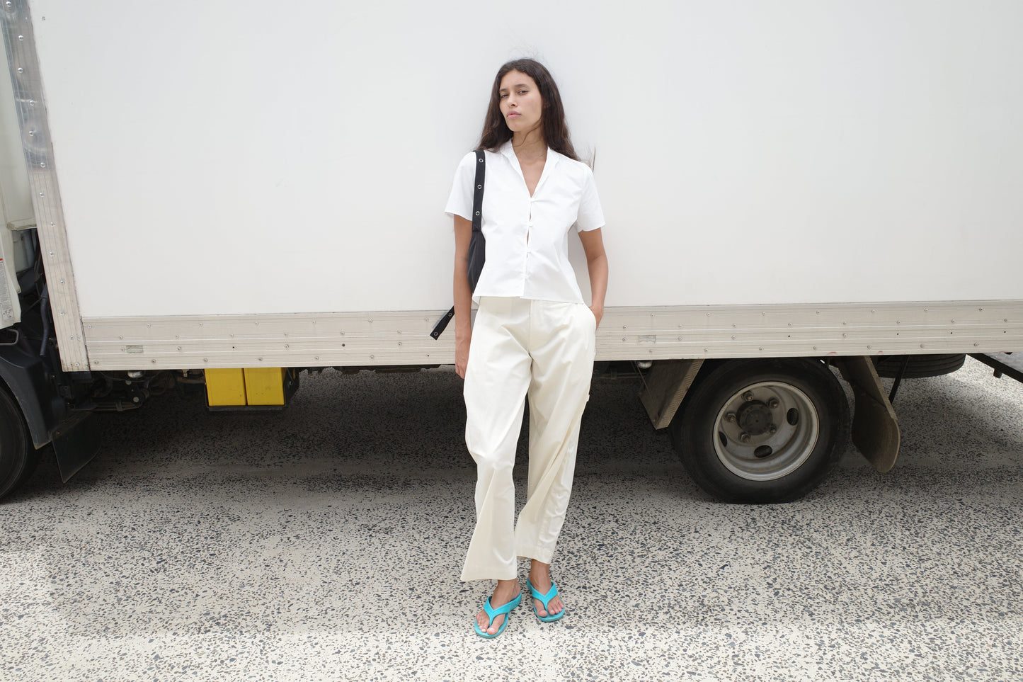 A woman in the Sails Top - White and loose white pants stands by a white truck, one hand in her pocket, looking to the side. She has long dark hair, turquoise flip-flops, and carries a black shoulder bag.